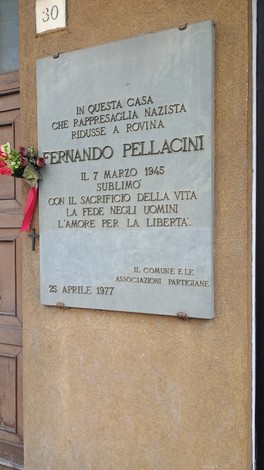 Lapide commemorativa dedicata a Fernando Pellacini, affissa su una parete esterna, con fiori decorativi.
