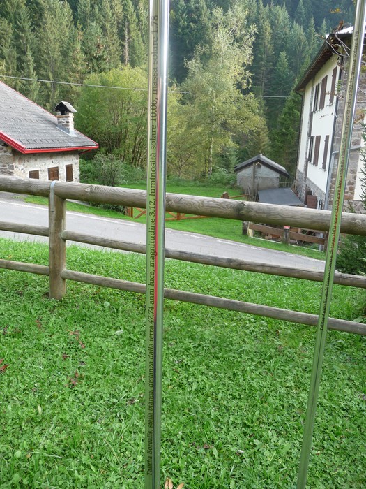 Monumento ai Dodici Caduti circondato da vegetazione e edifici in un paesaggio montano tranquillo.