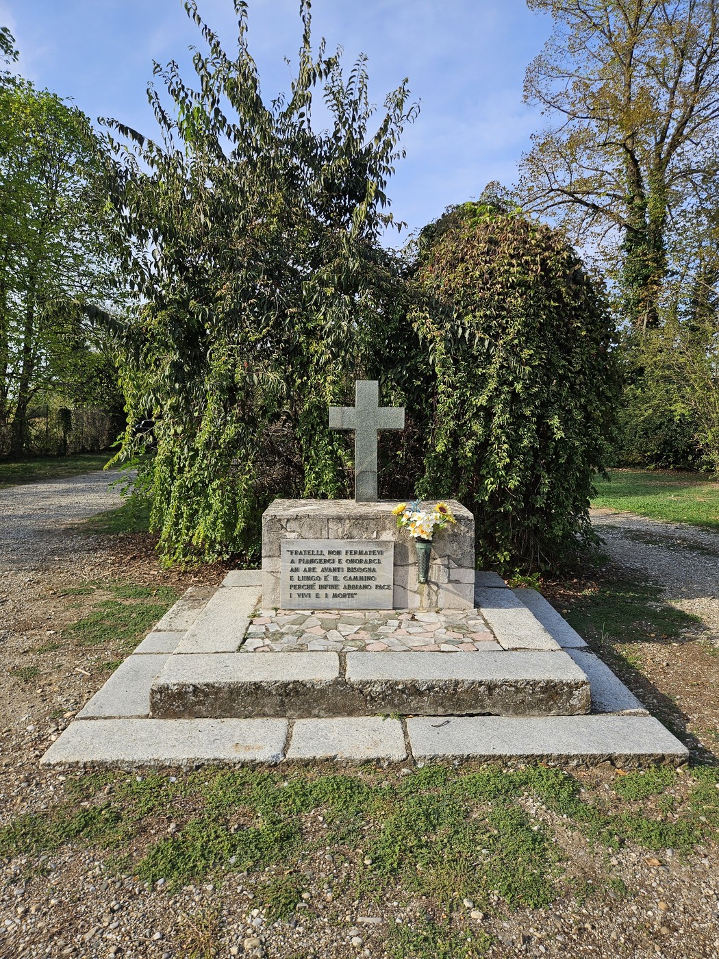 Monumento in pietra con croce, lapide con iscrizione e fiori, circondato da vegetazione in un parco.