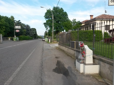 Cippo commemorativo dedicato a Bernini, situato lungo una strada tranquilla, con fiori in omaggio.