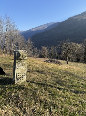 Cippo in pietra con iscrizione, circondato da un paesaggio montano e vegetazione armoniosa.