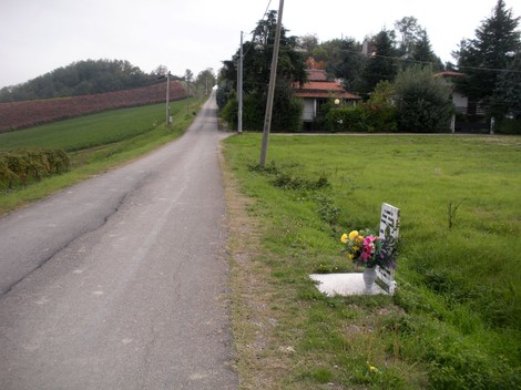 Lapide commemorativa in un paesaggio rurale, con strada e vigneti sullo sfondo. Fiori decorativi ai piedi della lapide.