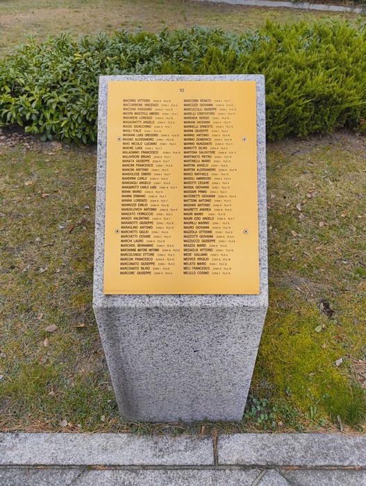 Targa commemorativa con nomi di caduti nel Cimitero militare italiano di Zehlendorf, circondata da vegetazione.