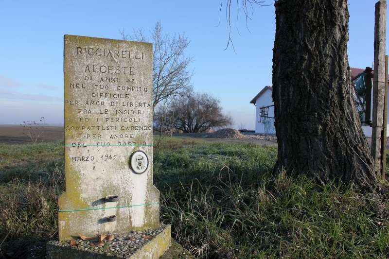 Cippo commemorativo a Ricciarelli, con incisioni e un ambiente rurale circostante.