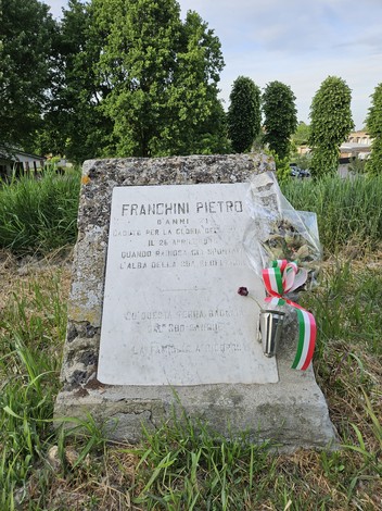 Cippo commemorativo a Franchini Pietro, con fiori e nastri tricolori, immerso nel verde di un campo.