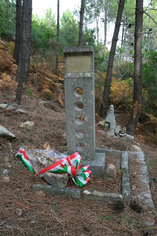 Cippo commemorativo in un bosco, adornato con una corona di fiori e simboli nazionali.