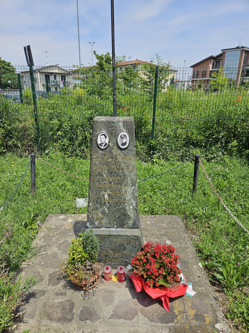 Cippo commemorativo con cerchi fioriti e fotografie, immerso nel verde di un'area urbana.