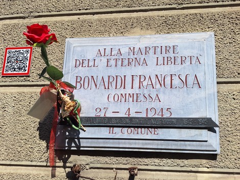 Lapide in marmo su parete con iscrizione, una rosa rossa e un mazzo di fiori appesi sotto.