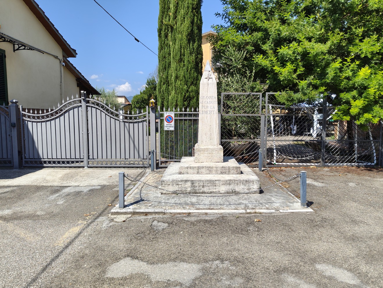 Cippo a forma di obelisco in pietra con iscrizione, su basamento a gradoni, in un cortile recintato.