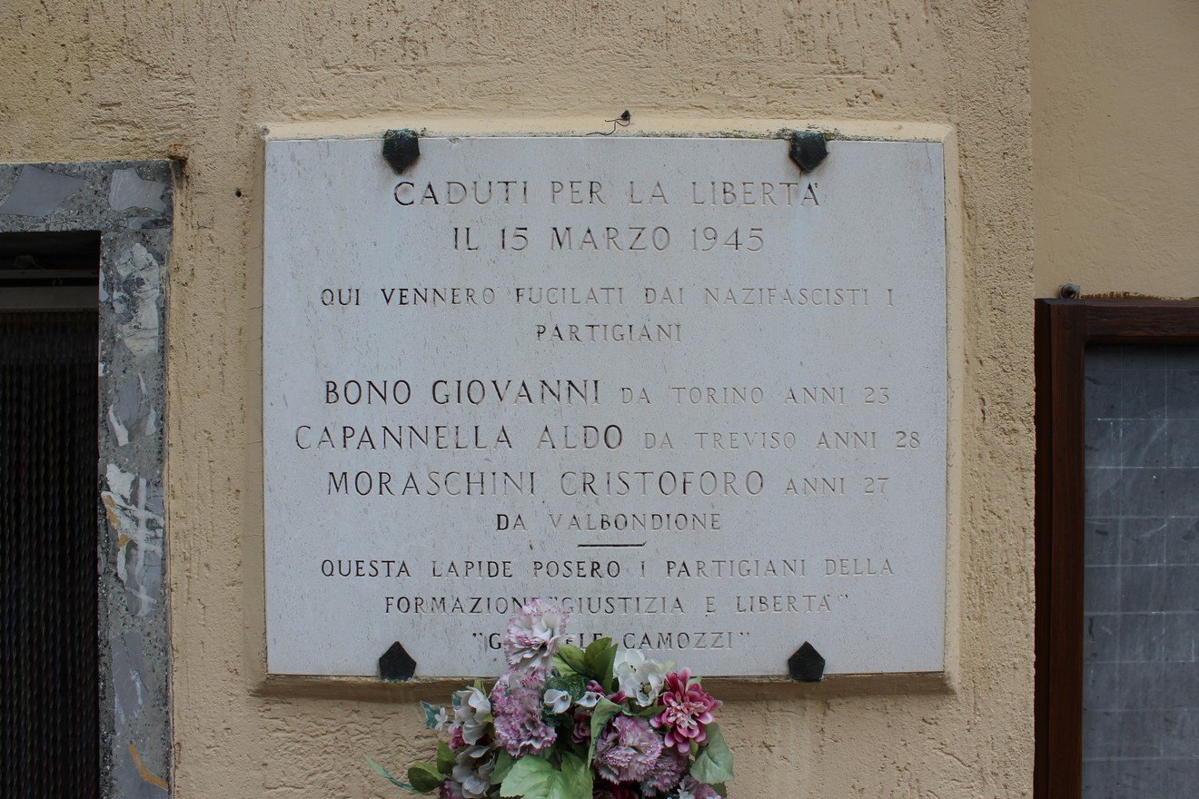 Lapide commemorativa in onore dei Martiri della Libertà, decorata con fiori, situata su un muro.