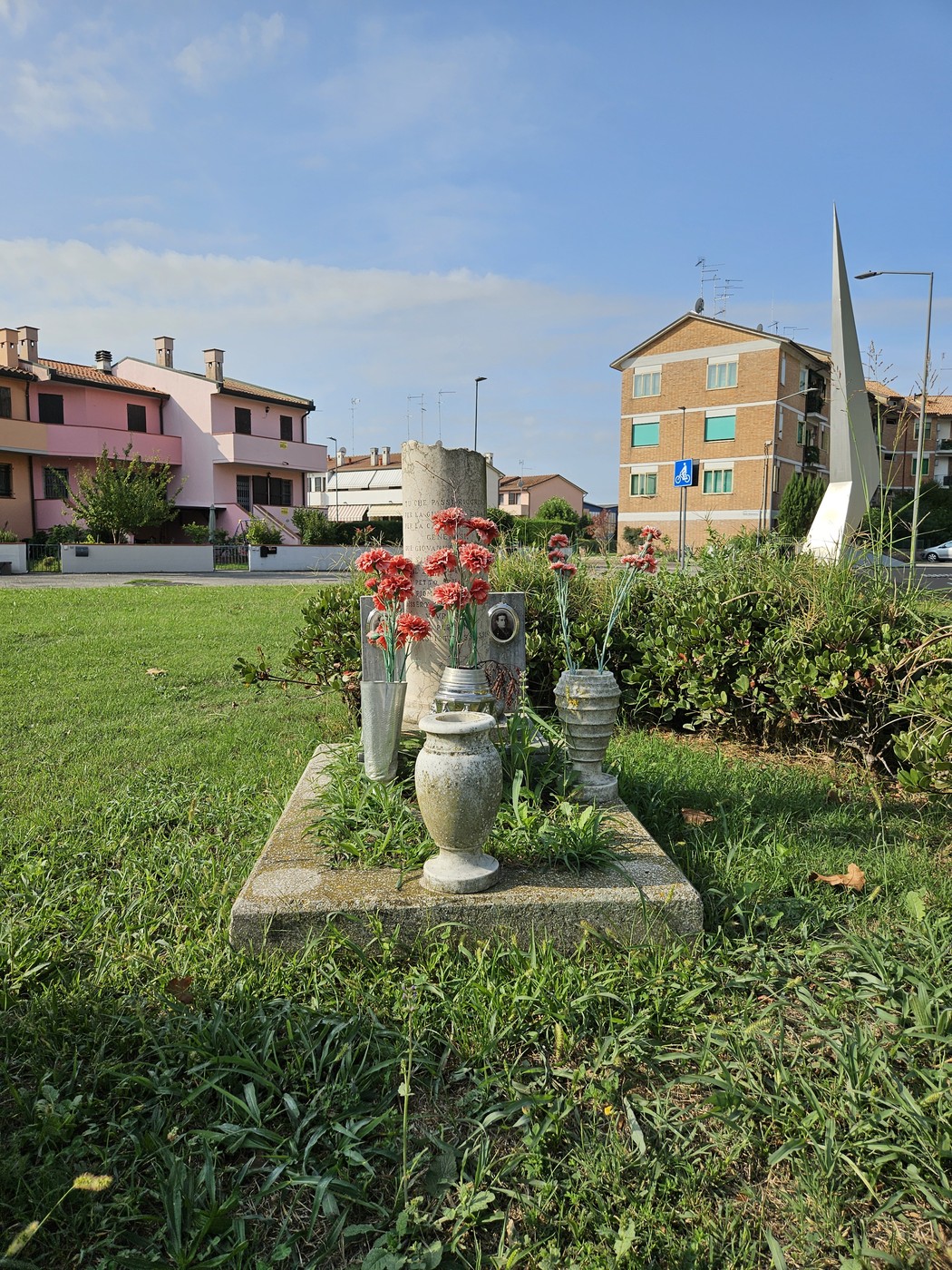 Cippo commemorativo con fiori e decorazioni, situato in un'area verde urbana. Edifici sullo sfondo.