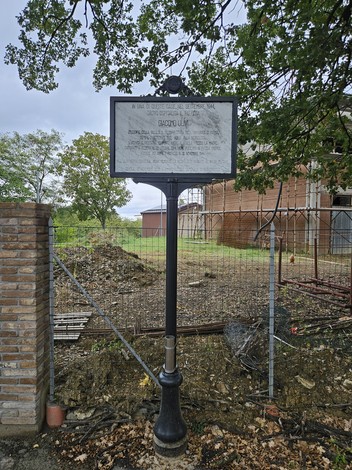 Lapide commemorativa circondata da verde, con sfondo di edificio e recinzione. Ambiente rurale e storicità.