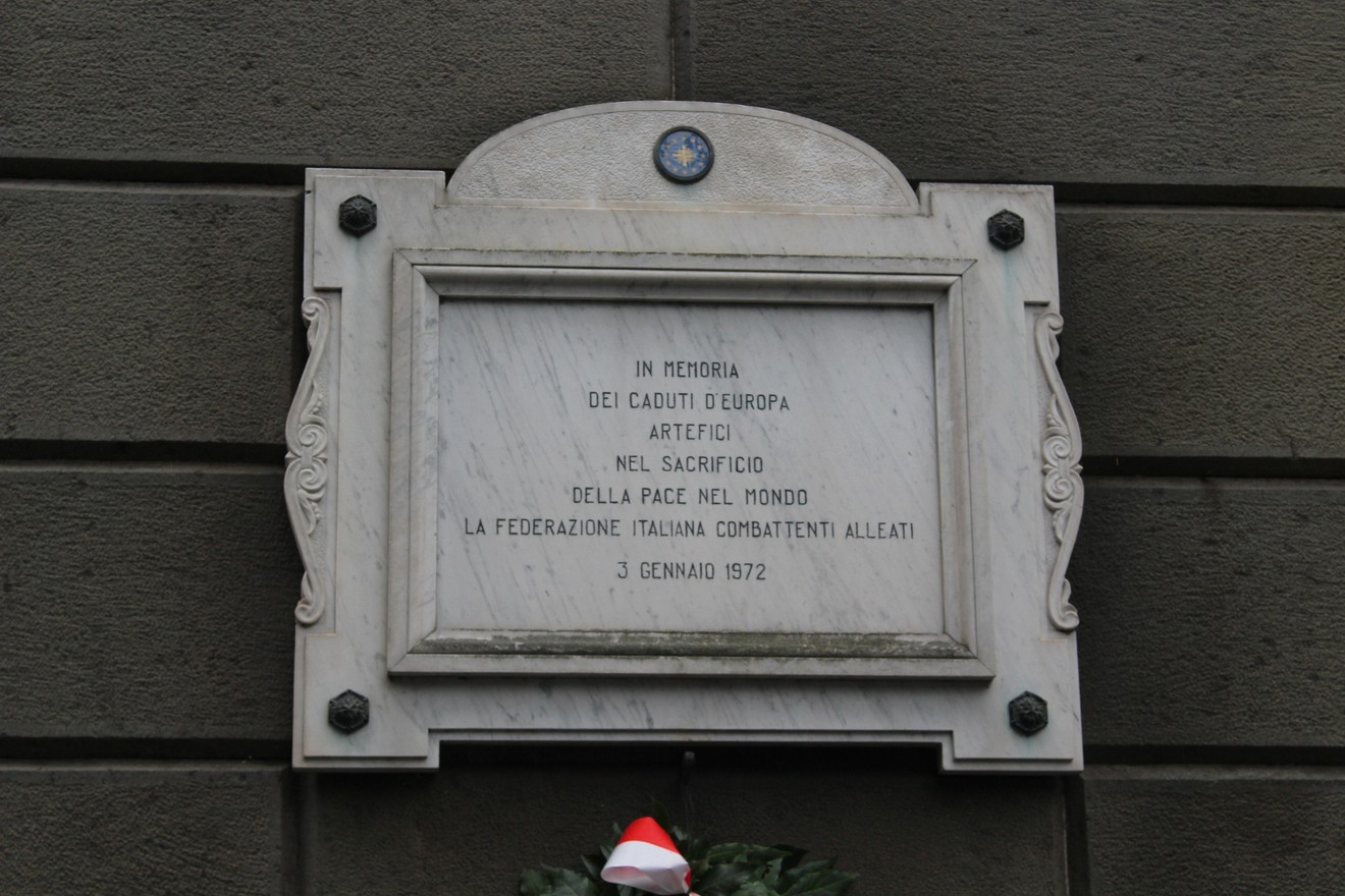 Lapide commemorativa dedicata agli Alleati caduti, situata in una parete esterna, con decorazioni floreali.
