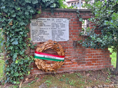Due lapidi con iscrizione su muro in mattoni, corona con nastro tricolore appoggiata sotto, edera intorno.