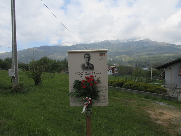 Targa commemorativa ad Aldo Galiazzo, circondata da un paesaggio montano e vegetazione verde.