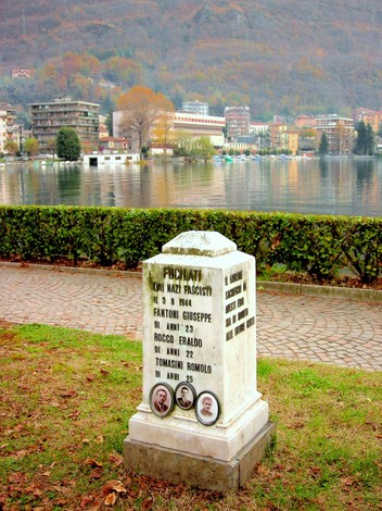Cippo commemorativo dedicato a Fantoni, Rocco e Tomasini, sul lungolago in un contesto naturale.