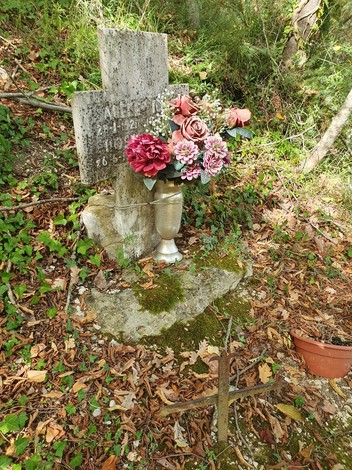 Croce commemorativa con fiori artificiali in un contesto naturale, circondata da foglie e vegetazione secca.