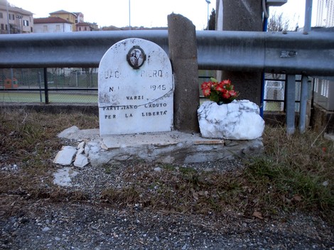Lapide commemorativa dedicata a un caduto per la libertà a Varzi, risalente al 1945. Fiori e pietre adornano il monumento.