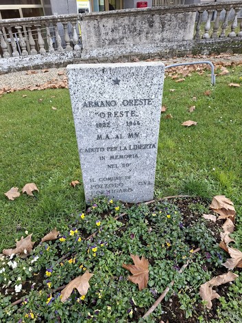 Cippo commemorativo dedicato a Armano Oreste, con dedica alla liberazione, circondato da fiori e foglie secche.