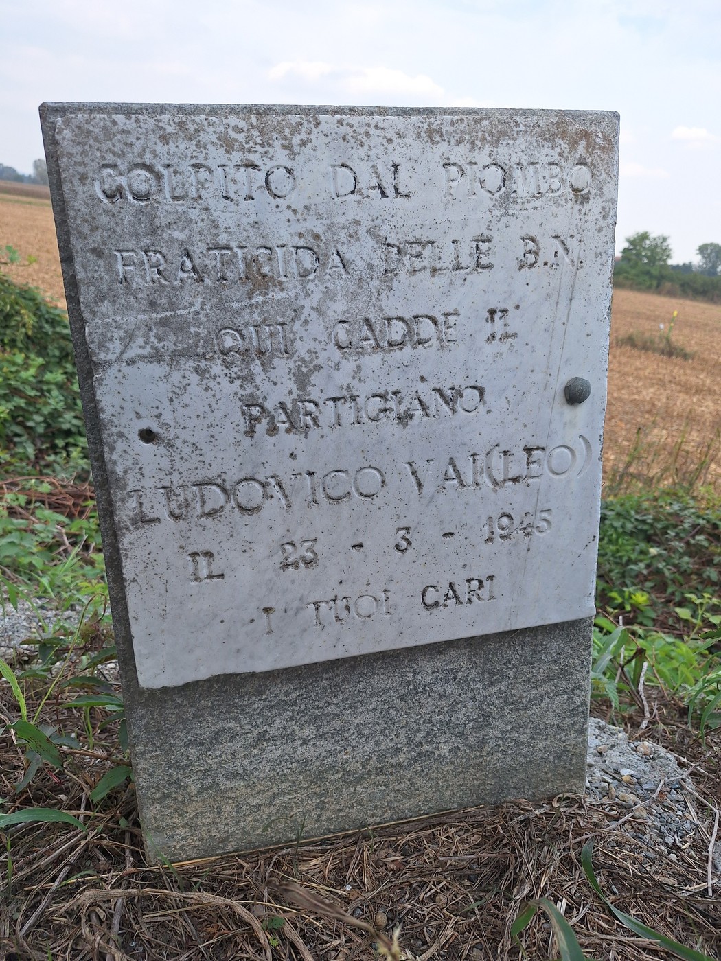 Cippo in pietra con iscrizione, in un campo erboso ai margini di una strada di campagna.