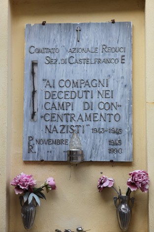 Lapide commemorativa dedicata ai caduti nei campi di concentramento nazisti, con fiori artificiali ai lati.