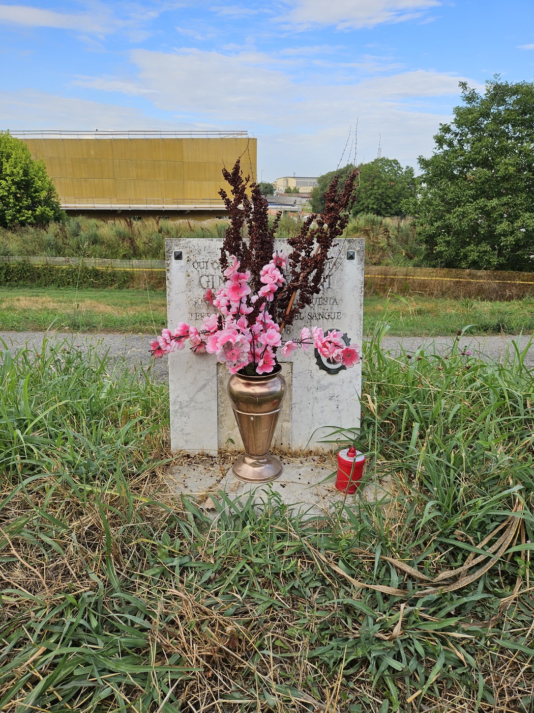 Cippo commemorativo con fiori e candele, circondato da erba e vegetazione, in una strada di Avigni.