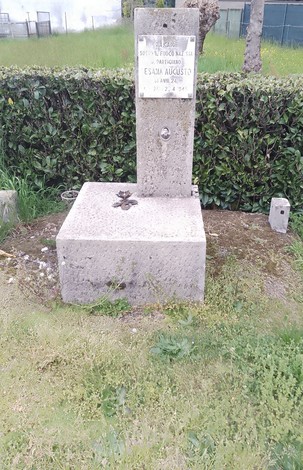Cippo commemorativo a Sesana, situato in un'area verde, circondato da siepi e con terreno erboso.