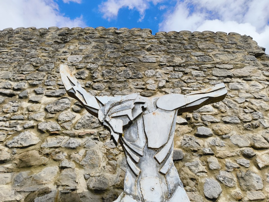 Scultura in metallo di un toro davanti alle storiche Rovine di Caprara di Sopra, con cielo blu e nuvole.