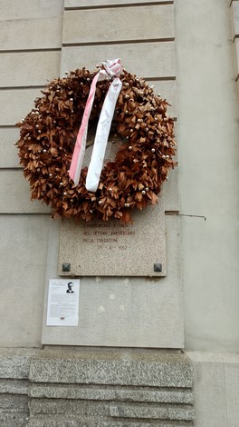 Lapide commemorativa con una corona di fiori secchi e nastri, apposta su una parete di pietra.