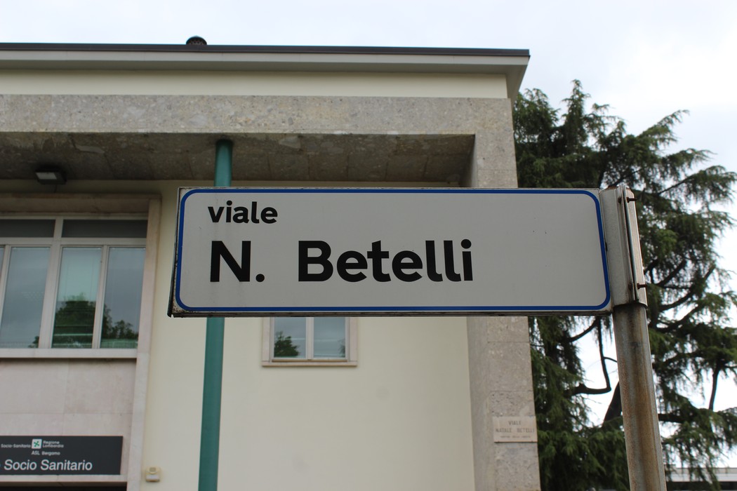 Targa stradale che indica viale N. Betelli, con un edificio sullo sfondo e alberi ai lati.