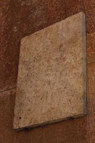Lapide in pietra affissa su muro, con iscrizioni visibili in parte, su sfondo di superficie ruvida.