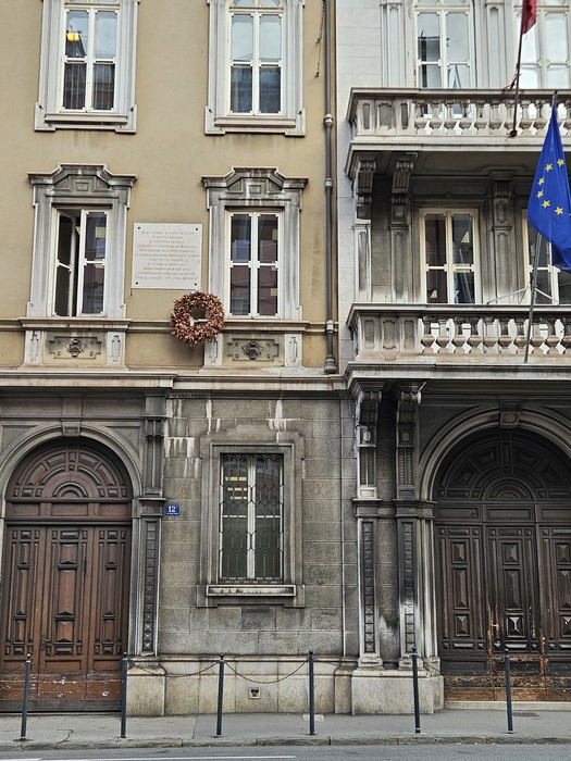 Lapide commemorativa affissa su un edificio, con decorazioni floreali e bandiere europee e italiane.