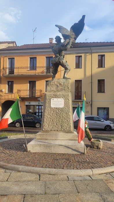 Monumento in bronzo e pietra con figura alata su piedistallo, al centro di una piazza, con bandiere italiane ai lati.