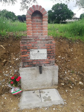 Tabernacolo dedicato a Michelucci, con struttura in mattoni e fiori rossi nel terreno circostante.