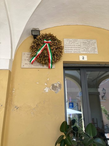 Lapidi commemorative all'esterno del Caffè Roma, con una corona di fiori e decorazioni tricolori.