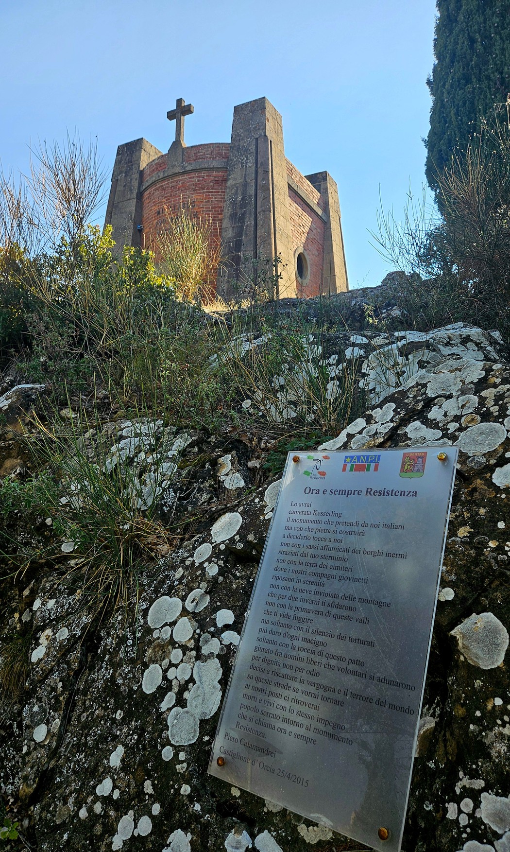 Targa commemorativa su roccia lichenata, in primo piano rispetto a una torre con croce sulla cima.