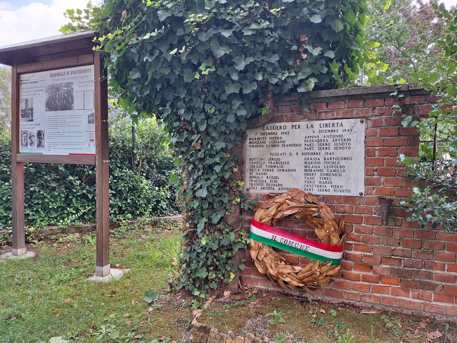 Lapide in marmo con iscrizione su muro di mattoni, corona tricolore appesa e edera sul muro retrostante.