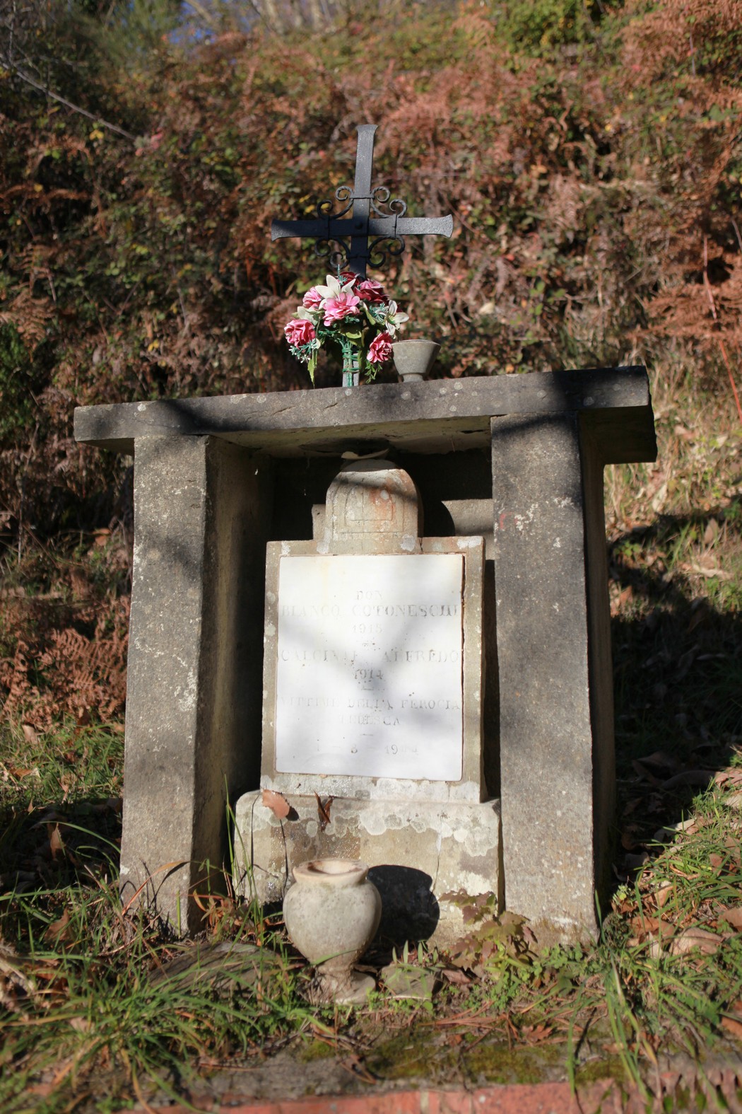 Cippo commemorativo con croce, fiori e vasi in un contesto naturale suggestivo.