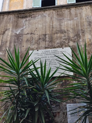 Lapide commemorativa per il genocidio dei nomadi, con vegetazione circostante e edificio storico sullo sfondo.