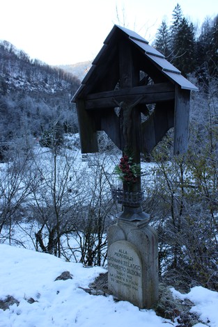 Croce in legno con statue a Duzioni, circondata da neve e alberi, in un paesaggio montano suggestivo.