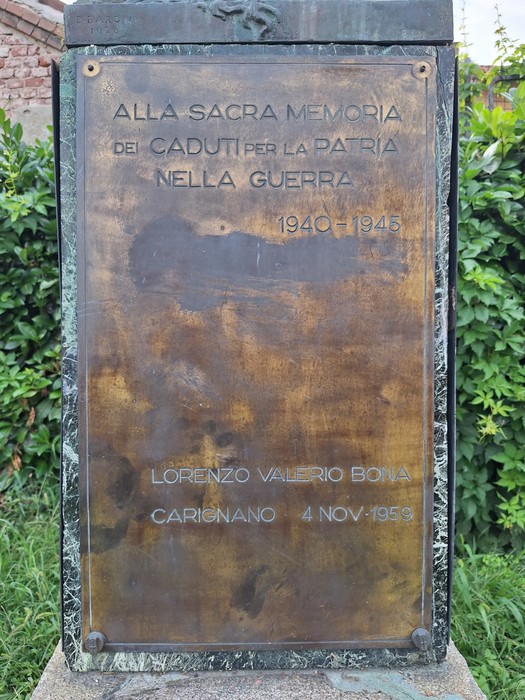 Targa in bronzo con iscrizione, montata su un cippo in pietra all'aperto, circondata da vegetazione.