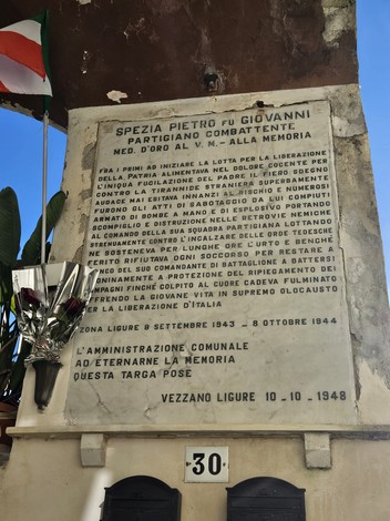 Lapide commemorativa dedicata a Pietro Giovanni, partigiano, a Vezzano Ligure, con fiori e bandiera italiana.