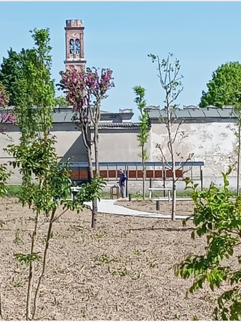 Giardino della Memoria con un sentiero curvilineo e alberi, sullo sfondo una torre e un edificio.