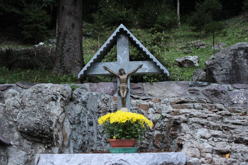 Monumento commemorativo con croce e statua di Cristo, circondato da fiori e un paesaggio naturale.