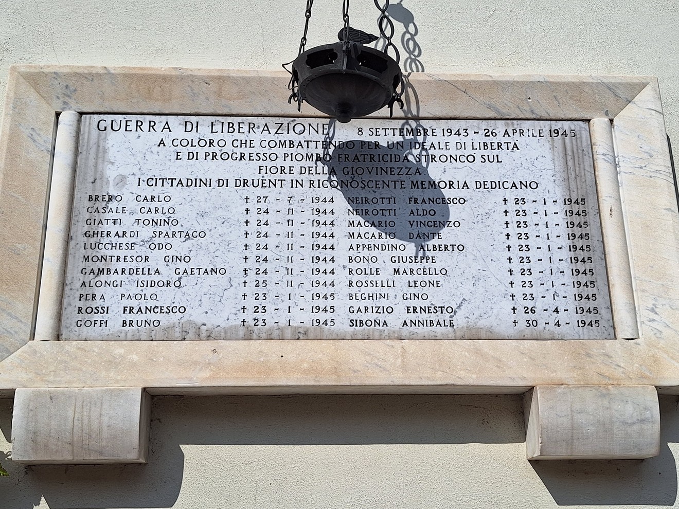 Lapide in marmo con lungo elenco di nomi e date, fissata su parete esterna, sormontata da lanterna.