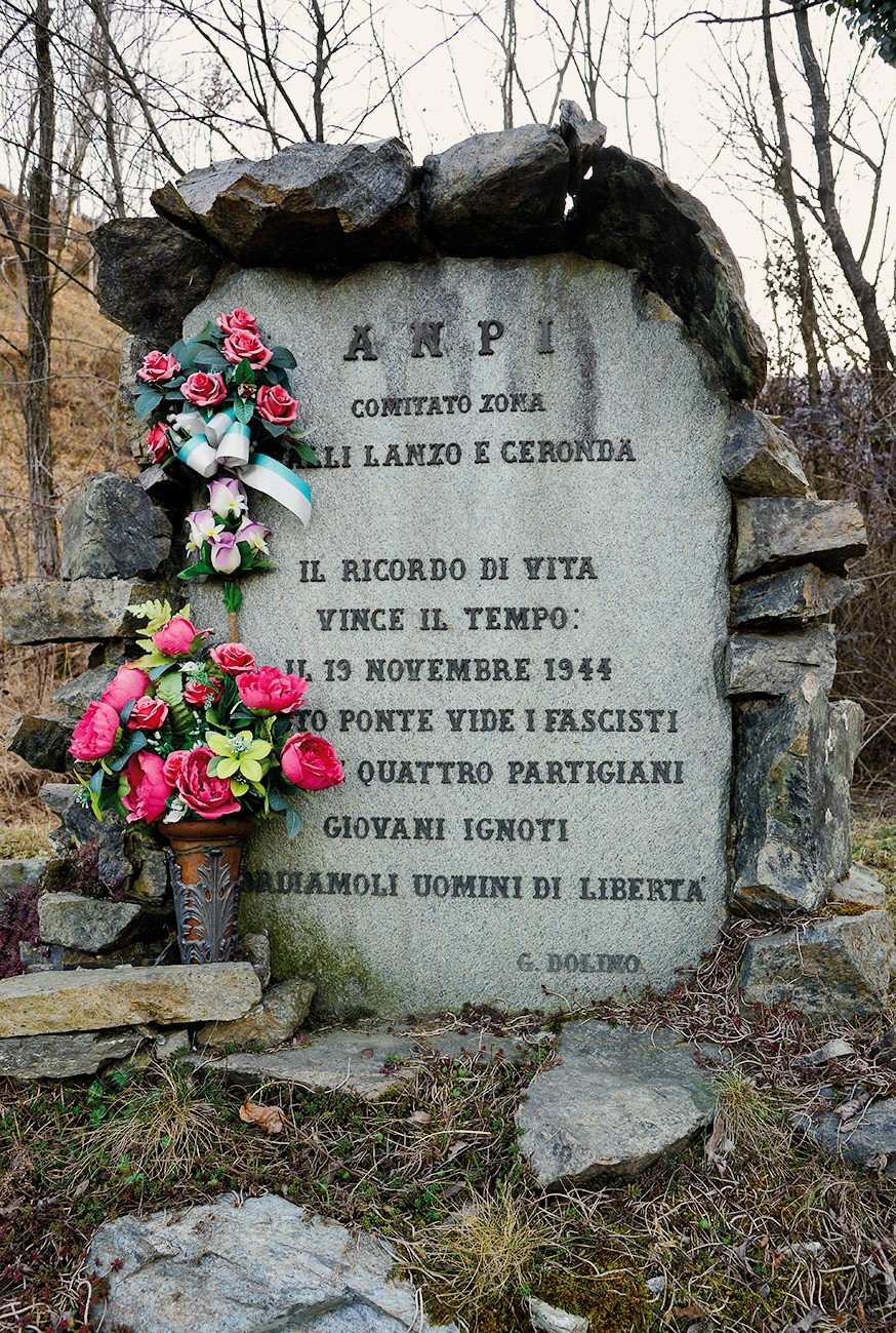 Cippo in pietra con iscrizione ANPI, incorniciato da massi, con mazzo di fiori ai piedi nel bosco.