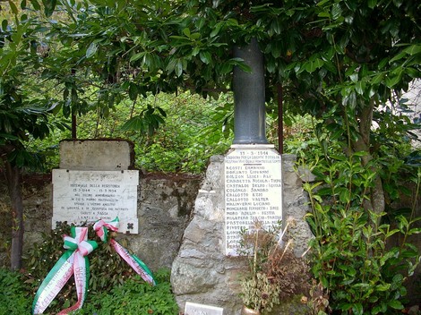 Lapide commemorativa dedicata ad Agosti e ad altri, circondata da vegetazione e decorazioni floreali.