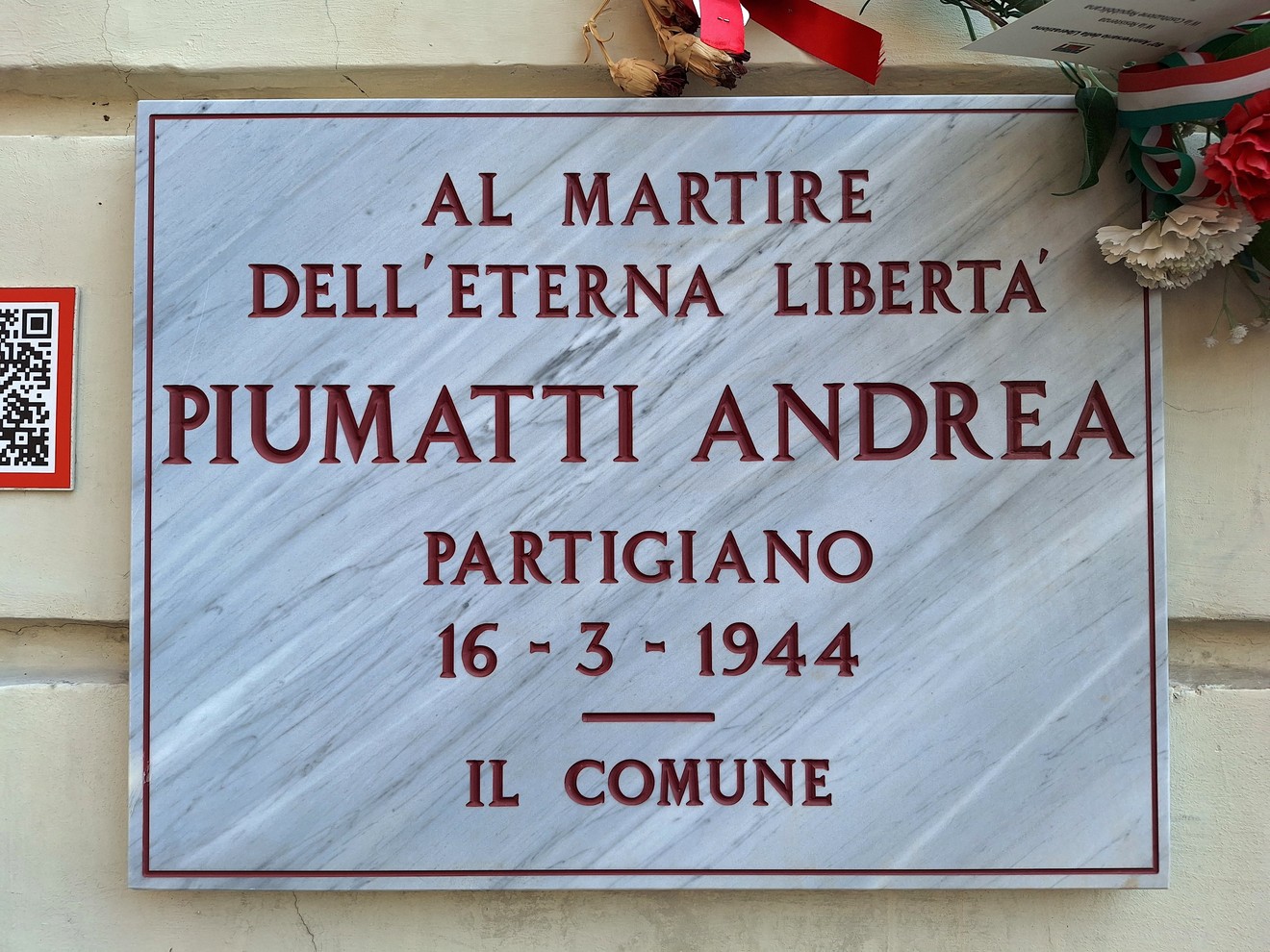 Lapide in marmo su parete, con iscrizione dedicata a un partigiano e fiori decorativi in alto.