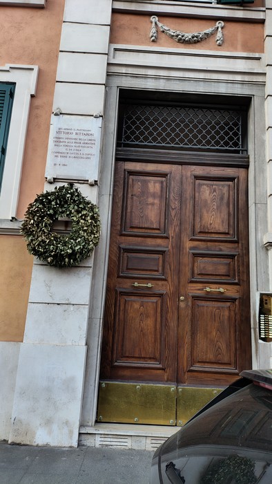 Lapide a Buttaroni accanto a una porta in legno con decorazioni vegetali. Ambiente urbano ben curato.