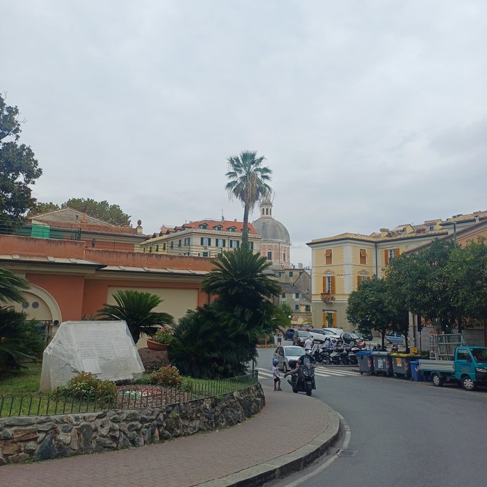 Lapide in pietra con iscrizione in un'aiuola di una piazza, tra palme e edifici d'epoca.